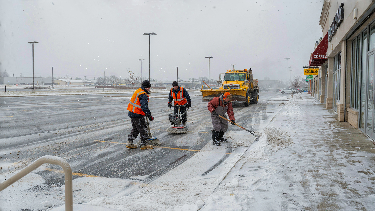 A sidewalk crew clears snow beside a commercial parking lot in daytime